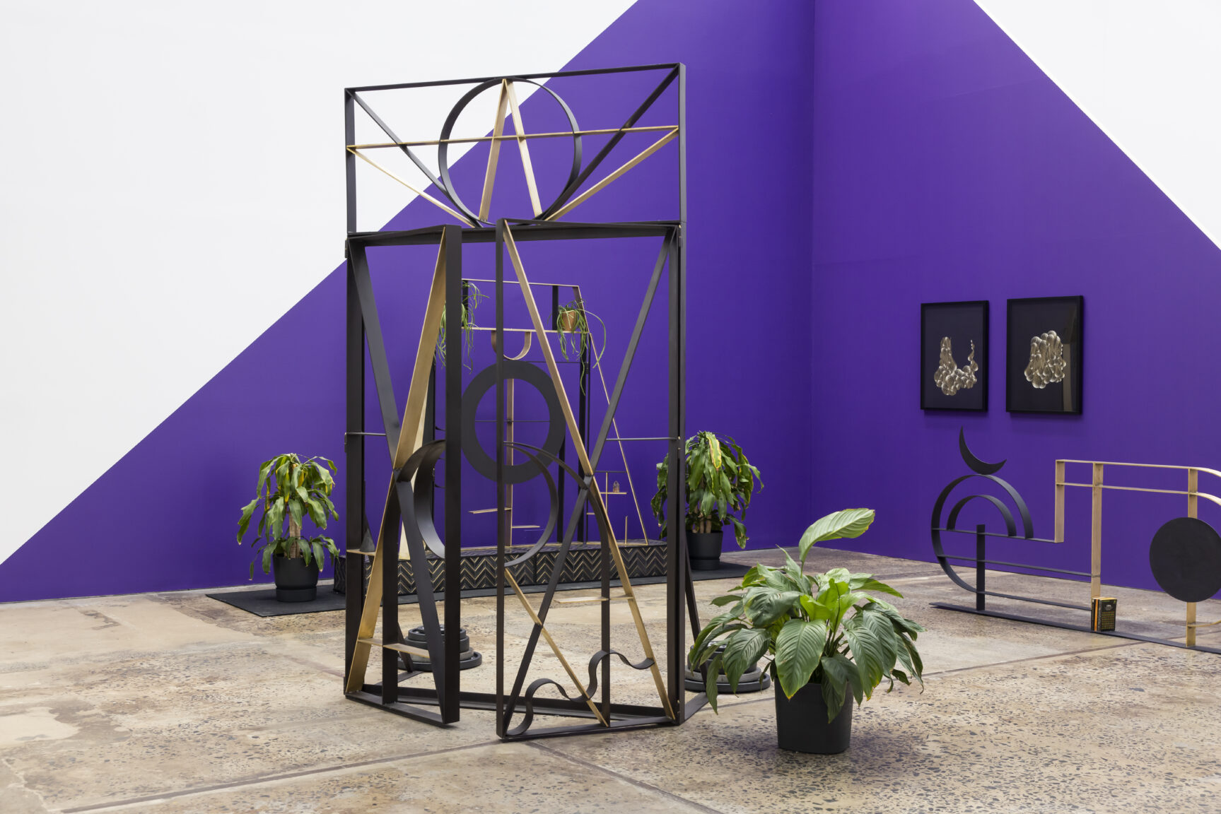 An art installation, inclusive of two wrought-iron like structures, three pot plants and two framed images hanging on a wall that forms a corner with another wall. The wall is painted diagonally in two-tone, white and purple with the purple sections creating a triangular like shape from the corner.