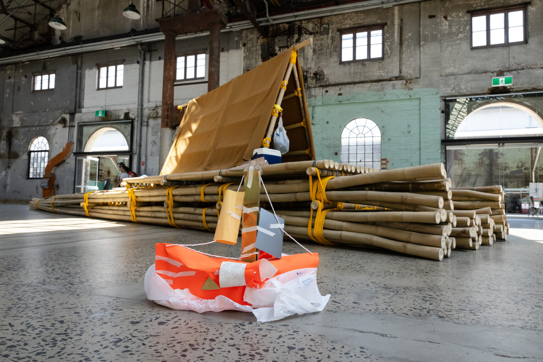a bamboo raft inside the Public Space of Carriageworks behind crafted cardboard recreation sitting of the bamboo