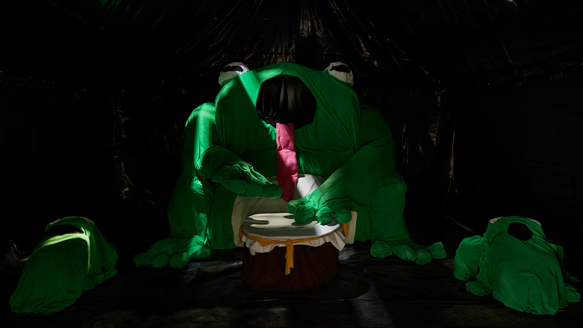 A large frog made of fabric sits in a large, dark room flanked by two smaller frogs.