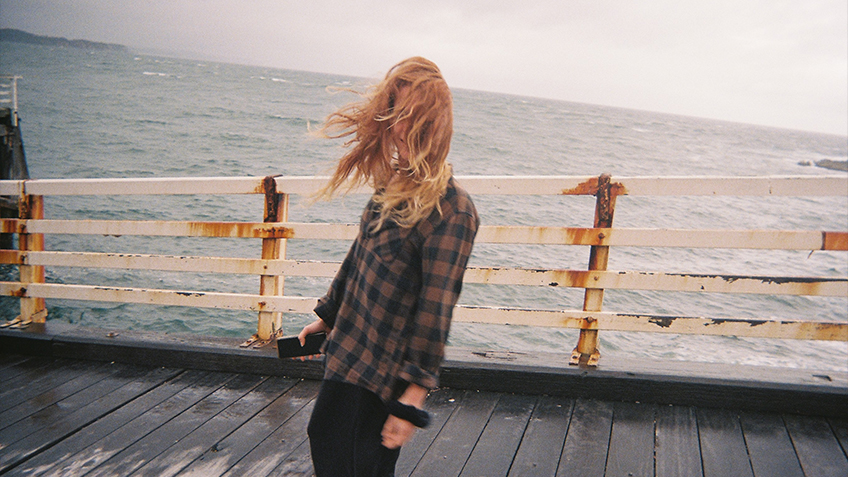 amby stands on a wet wooden jetty on an overcast day. A grey ocean stretches out behind them while the wind pushes their hair into their face, obscuring a smile.