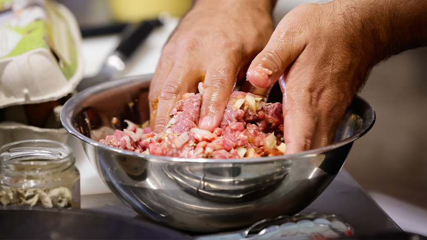 Two hands mix the ingredients for a burger patty in a silver bowl.