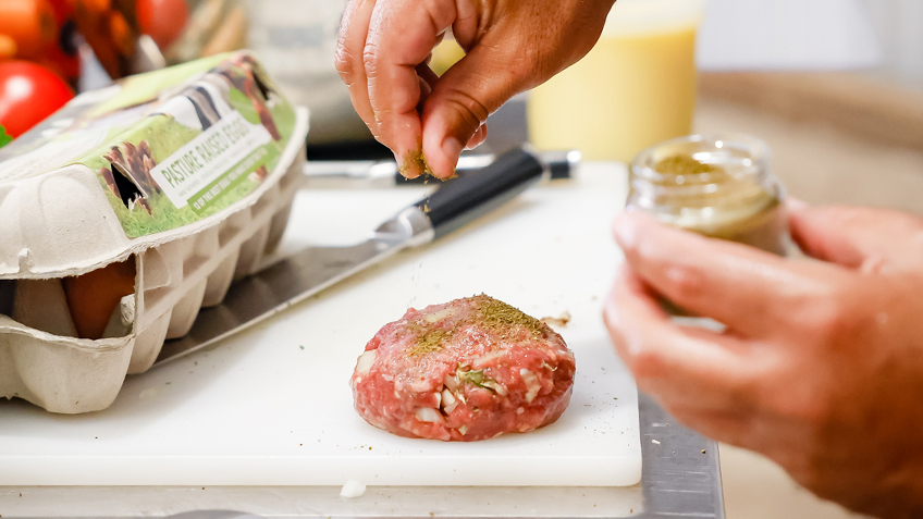 A hand seasons a burger patty on a white chopping board