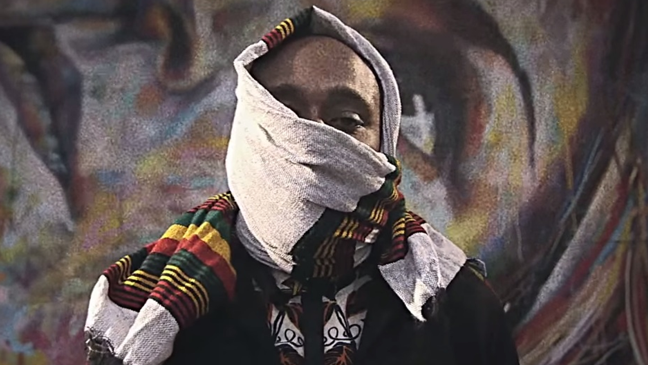 Yasiin Bey (Mos Def) seen from the shoulders up with a sweater wrapped around his face