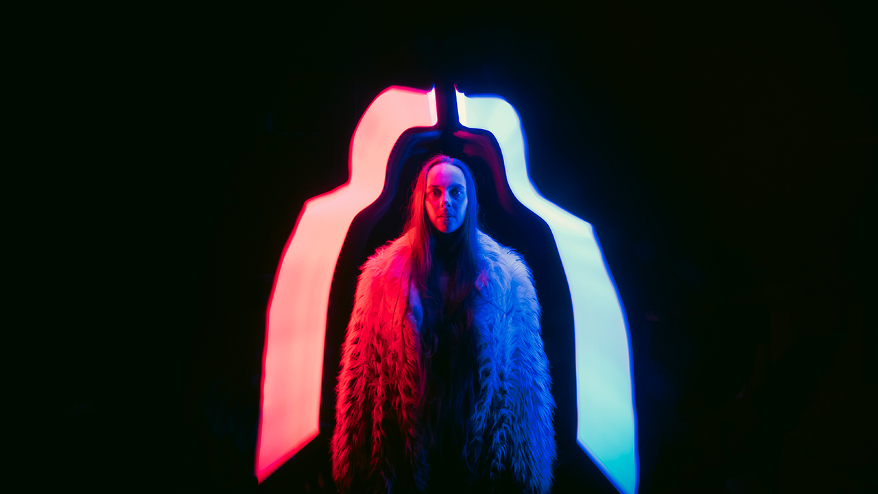 Chase stands in a fur coat underneath red and blue light against a black background.