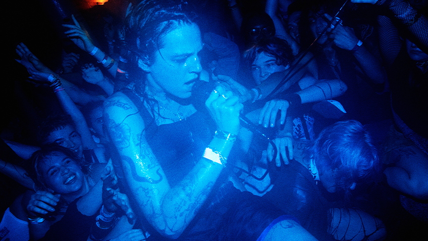 In a dark blue-lit room, deli girls' Tommi Kelly sings into a microphone while crowd surfing.