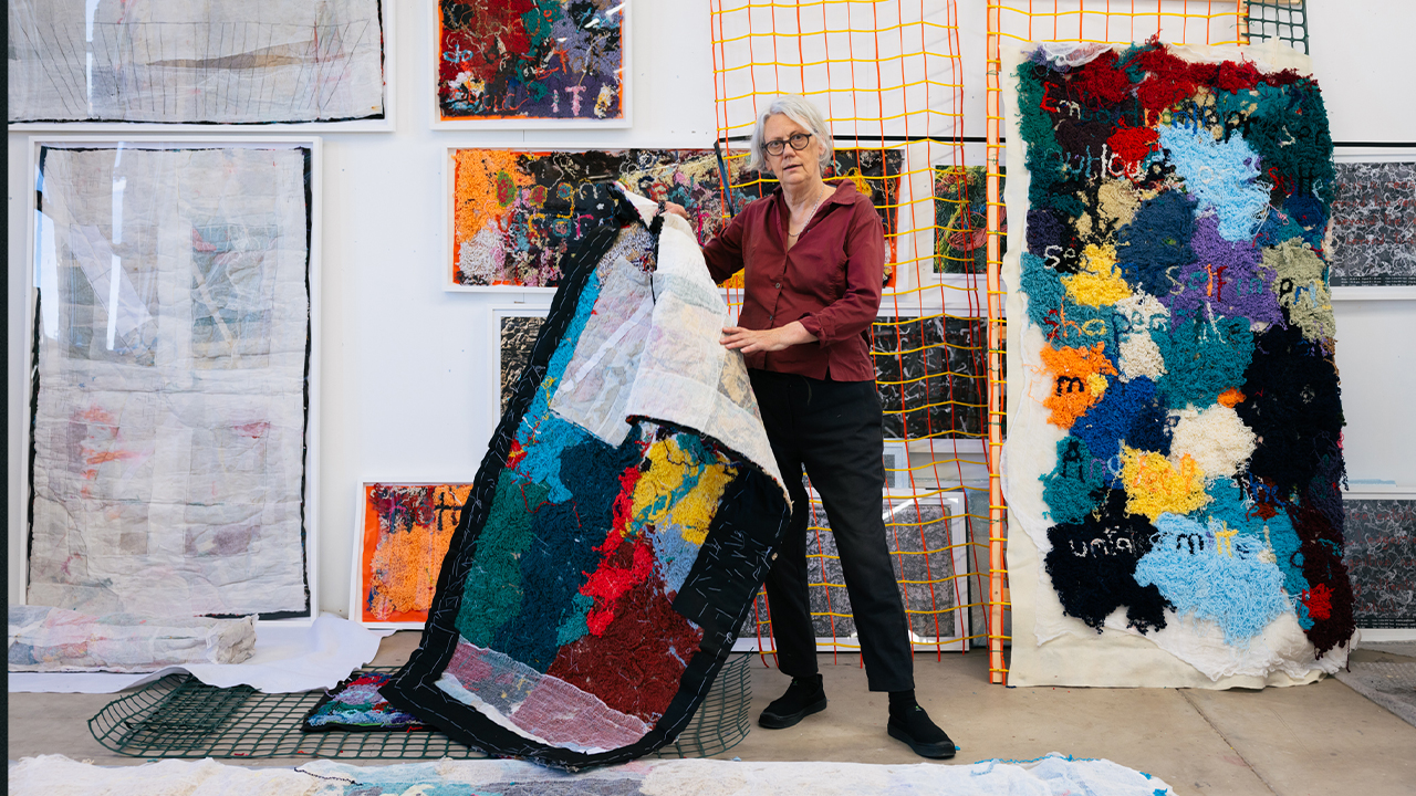 Artist Elizabeth Day stands in studio holding one piece of her stitched work.