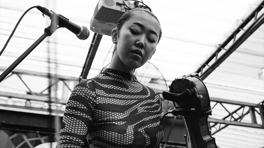 In a black and white photo, Elle Shimada stands at a deck holding a violin and wearing a patterned bodysuit.