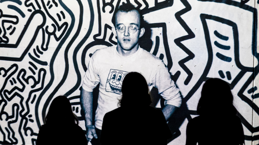 A photo taken from behind patrons watching a video tribute to artists lost to HIV/AIDS. Keith Haring is currently on screen.