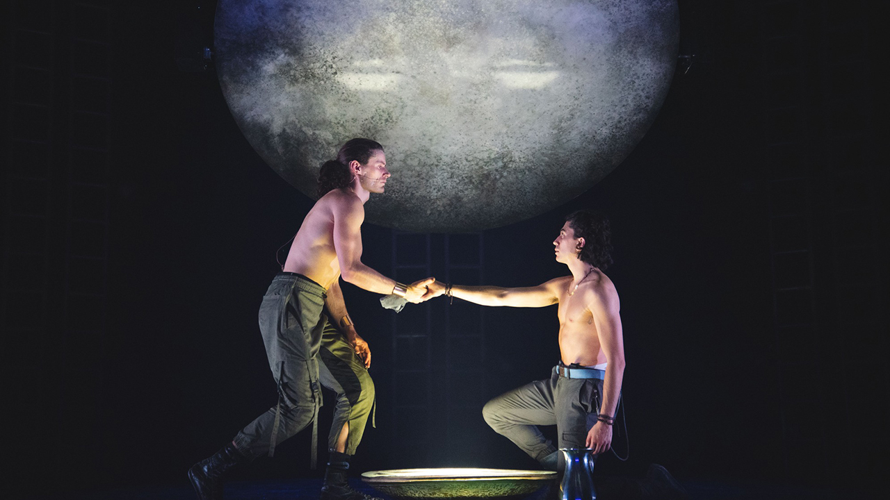 Two performers on stage hand-in-hand with a projection of the moon behind them.