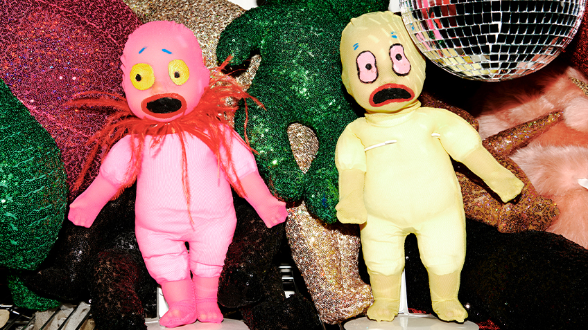 A pink and green doll sit on a shelf next to various glittery, sequined items.