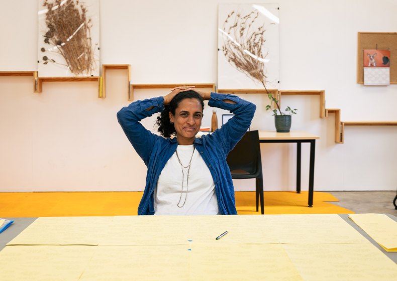 Keg De Souza sits back with her hands behind her head behind a table covered with yellow paper.