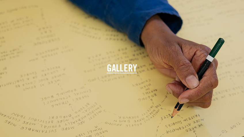Keg De Souza's hand holding a black pencil with a green tip over yellow paper. The text overlay reads 'Gallery'.