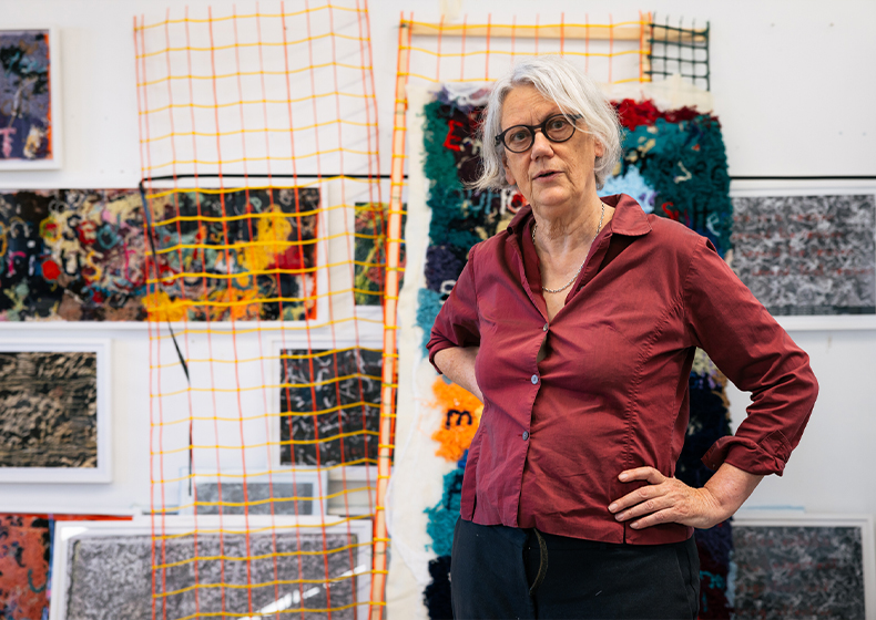 Liz Day stands with her hands on her hips in front of her artworks in her studio