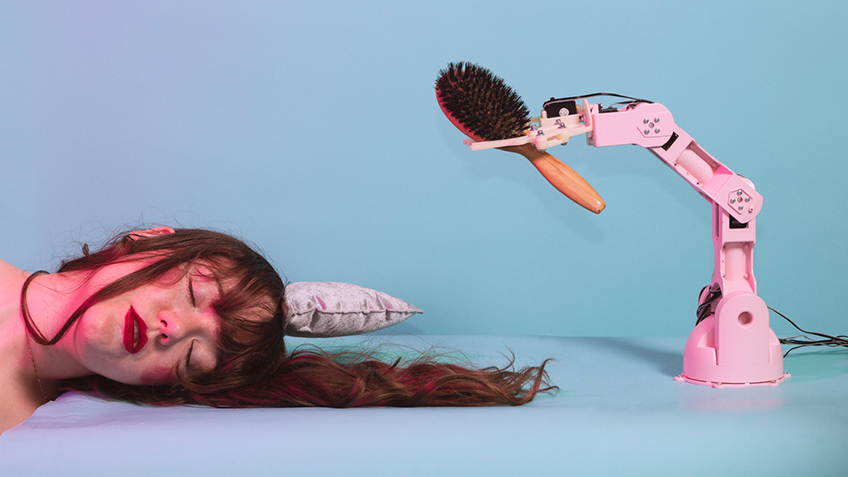 A woman's head lays on the ground with a pink robotic arm holding a brush on the other side.