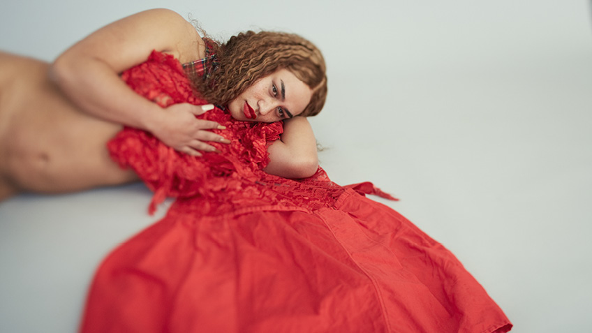 An artist lays on their side clutching a red dress to their chest