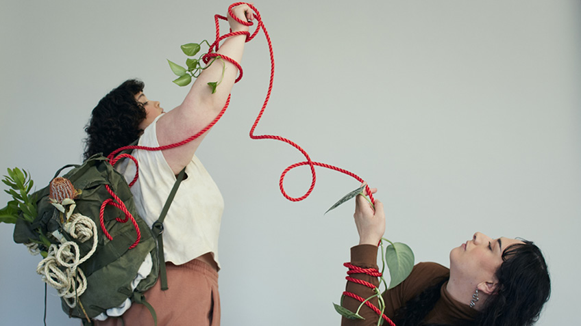 Two artists are wrapped together with red rope amongst green leaves.