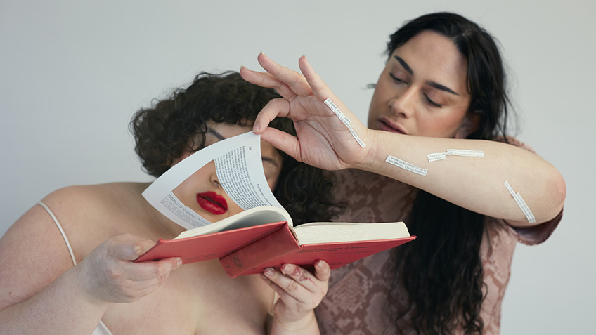 Two artists read a book, there is a square cut into the page and strips of text are stuck on the others arm.