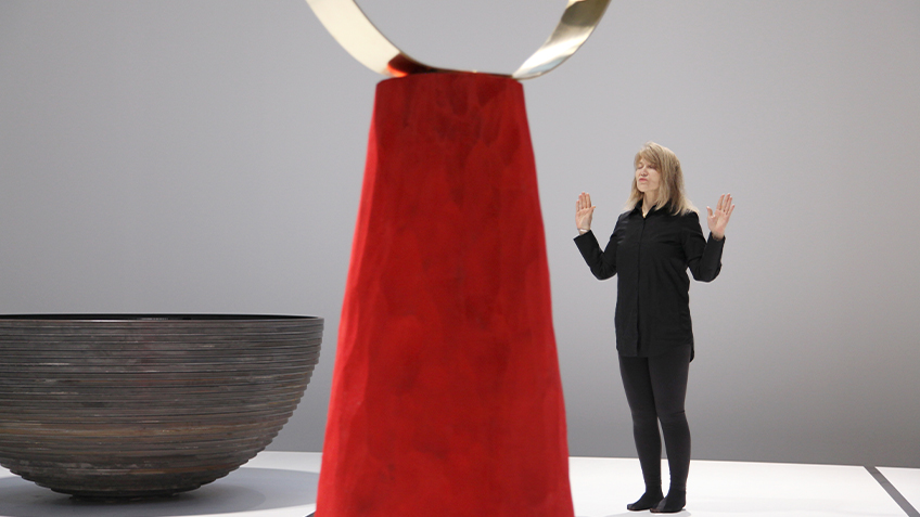 A woman dressed in black stands with her eyes closed and her hands up facing a red and gold sculpture. Next to the sculpture is a large brown bowl.