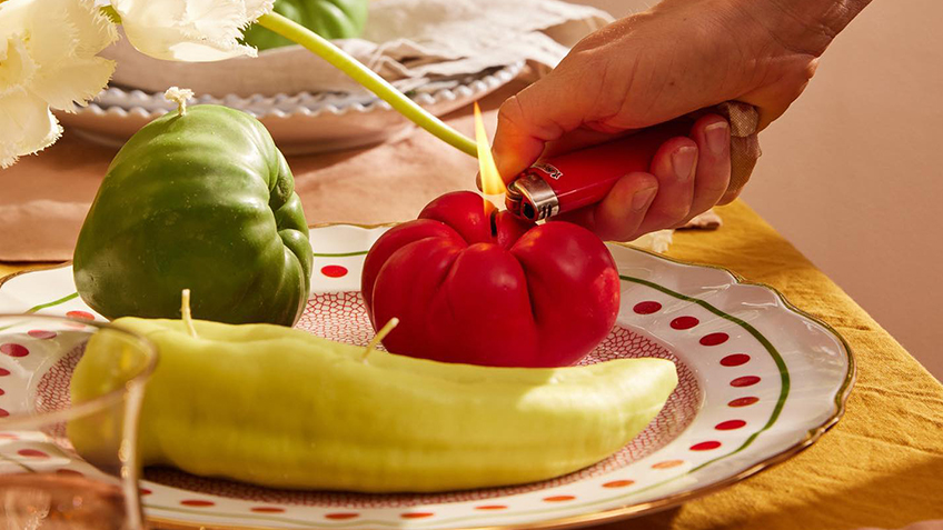 A hand lights one of three Nonna's Grocer candles on a plate on a yellow tablecloth