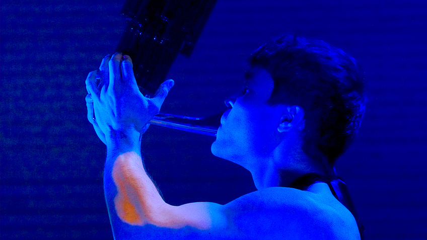 In deep blue light, Marcus Whale is shirtless playing a Sheng, an ancient mouth organ.