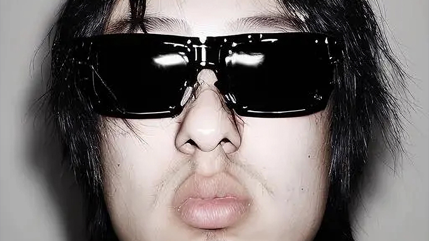 Against a grey background, Nerdie stares into the camera in a close up shot wearing black sunglasses.