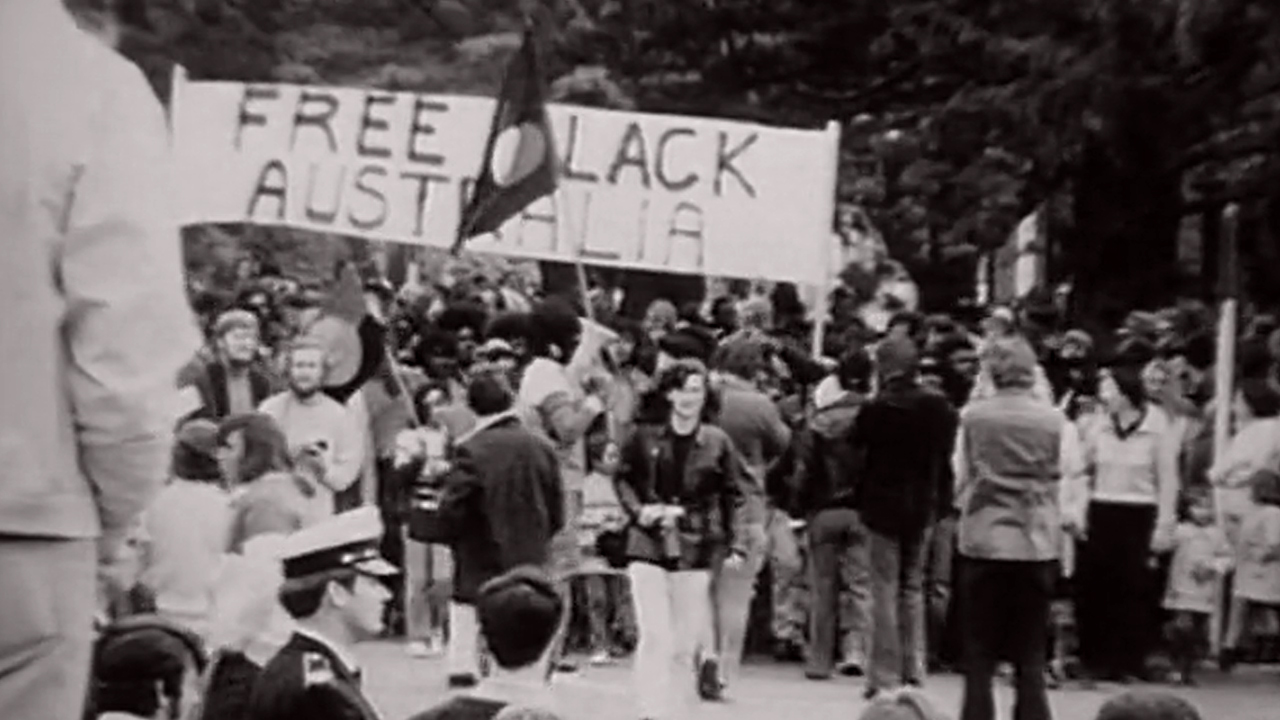 A black and white grainy image of a protest. Protesters hold a sign that reads 'FREE BLACK AUSTRALIA' - the letter B is covered by someone holding an Aboriginal flag.