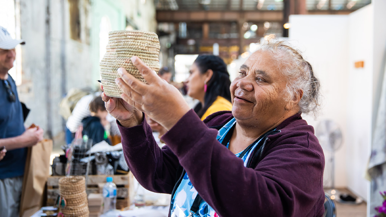 An artist from SOUTHEAST Aboriginal Arts Market holds up a woven basket.