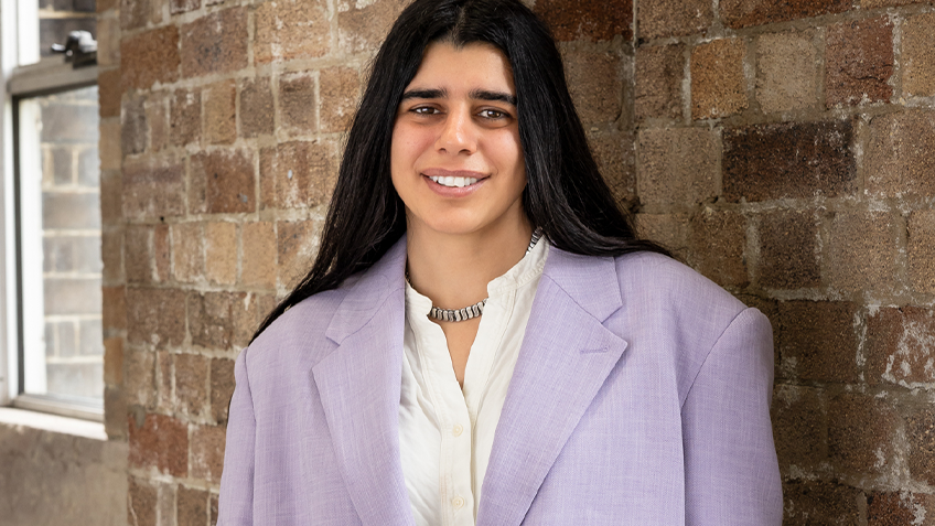 Shireen standing in front of a brick wall, wearing a lavender blazer and white button down shirt