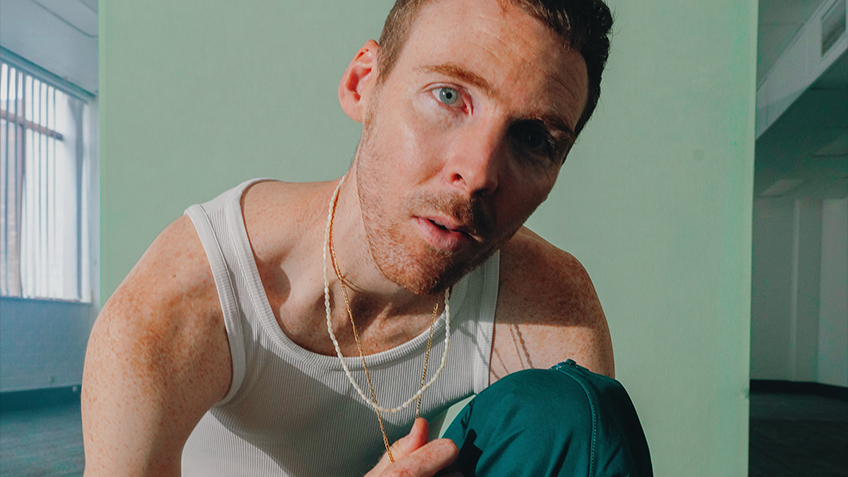 In a sterile office building, Skeleten kneels down to the camera wearing a white singlet and holding the gold chain around his neck.
