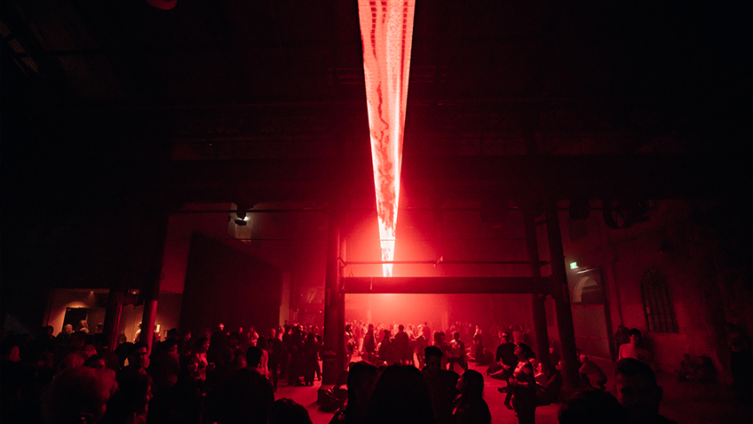 In a dark hazy room, a crowd socialises beneath a striking red light installation.