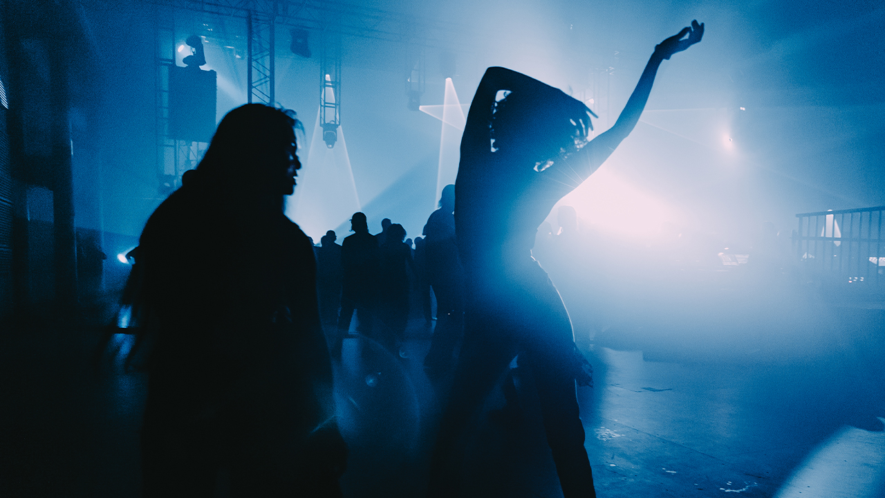 A silhouette of a person stands against blue light dancing with one arm in the air