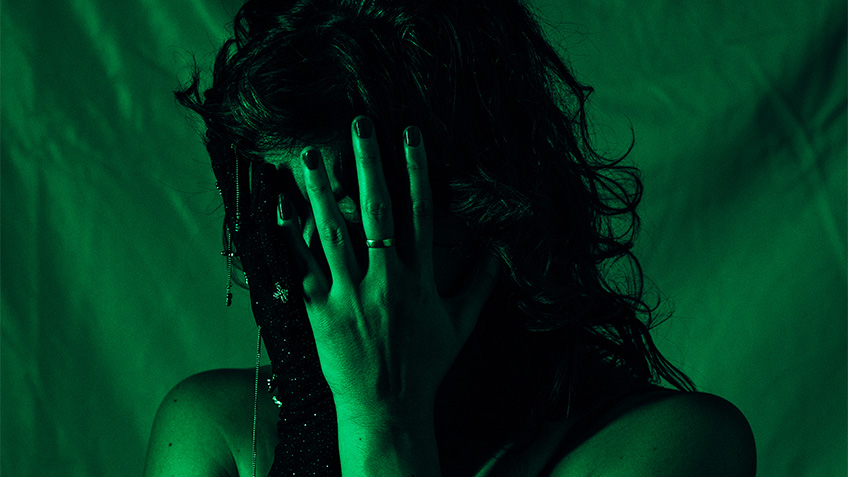 A women, bathed in green light covers her face with her left hand. She wears a wedding band on her ring finger.