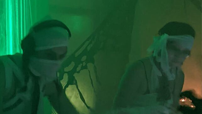 In a green-lit room, D-Grade and Wingnut are at decks wearing strips of white cloth wrapped around their faces and shoulders.