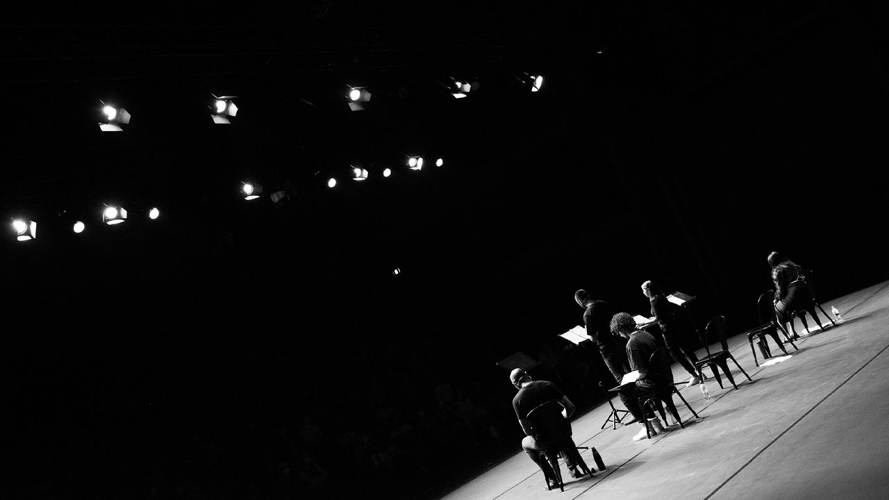 A black and white image of artists on stage taken from behind facing the audience.